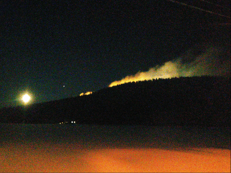 Mountain with snow floating from the snow guns, light up by the lights on the ski slopes against a dark sky.